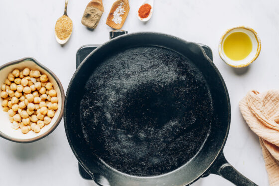 A cast iron skillet with various spices laid around it as well as a bowl of chickpeas.