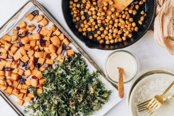 All the ingredients to build a vegan protein bowl displayed together. A sheet pan with roasted sweet potatoes, red onion and kale, a cast iron skilled with sautéed chick peas and a mason jar filled with creamy homemade white bean dressing.