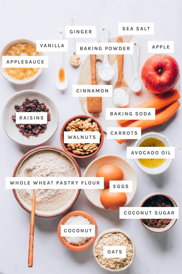 Ingredients measured out to make morning glory muffins: applesauce, vanilla, ginger, baking powder, sea salt, apple, cinnamon, baking soda, raisins, walnuts, carrots, avocado oil, whole wheat pastry flour, eggs, coconut sugar, coconut and oats.