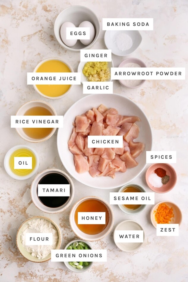 Ingredients measured out to make healthy orange chicken: eggs, baking soda, ginger, arrowroot powder, orange juice, garlic, rice vinegar, chicken, spices, oil, tamari, sesame oil, honey, water, orange zest, green onions and flour.