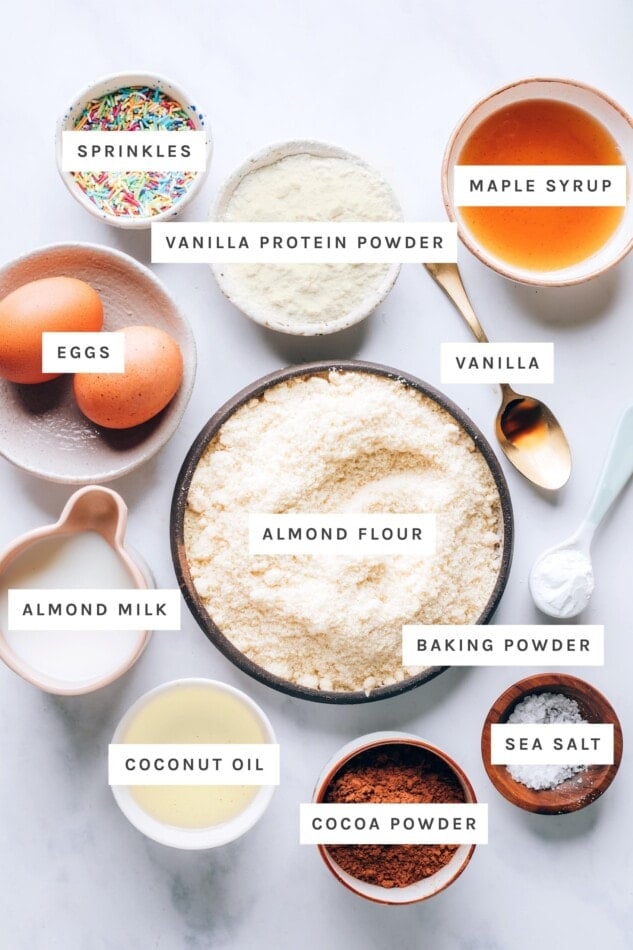 Ingredients measured out to make protein donuts: sprinkles, vanilla protein powder, maples syrup, eggs, vanilla, almond flour, almond milk, baking powder, coconut oil, sea salt and cocoa powder.