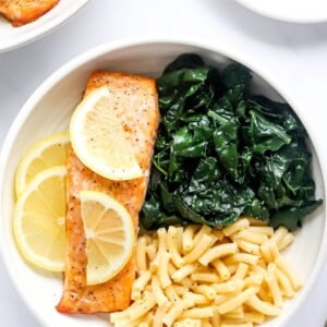A bowl with macaroni and cheese, kale and a filet of salmon. Lemon slices top the salmon filet.
