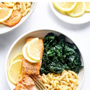 A bowl with macaroni and cheese, kale and a filet of salmon. A fork is breaking apart the salmon filet.