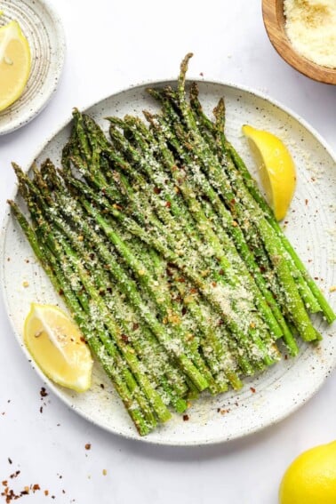 A plate with asparagus and lemon wedges topped with cheese and red pepper flakes.