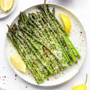 A plate with asparagus and some lemon wedges. The asparagus is topped with cheese and red pepper flakes.