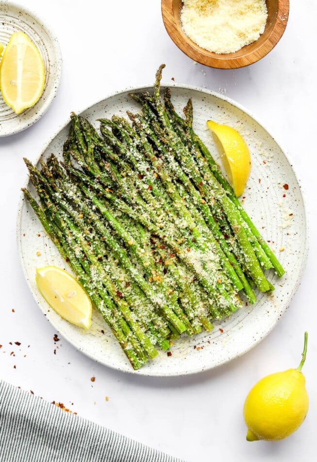 A plate with asparagus and some lemon wedges. The asparagus is topped with cheese and red pepper flakes.