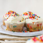 Four almond flour cupcakes wrapped in paper liners on a plate. The cupcakes have been topped with vanilla icing and rainbow sprinkles.