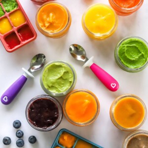 Ten varieties of baby food purees in jars with strawberries, blueberries, spoons and ice cube trays scattered around them.