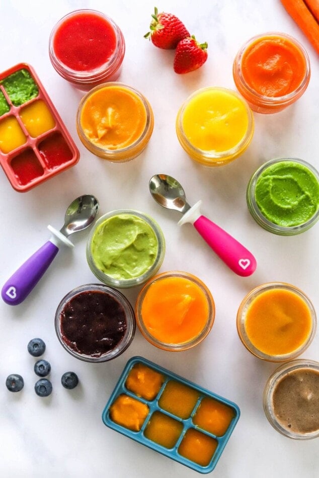 Ten varieties of baby food purees in jars with strawberries, blueberries, spoons and ice cube trays scattered around them.