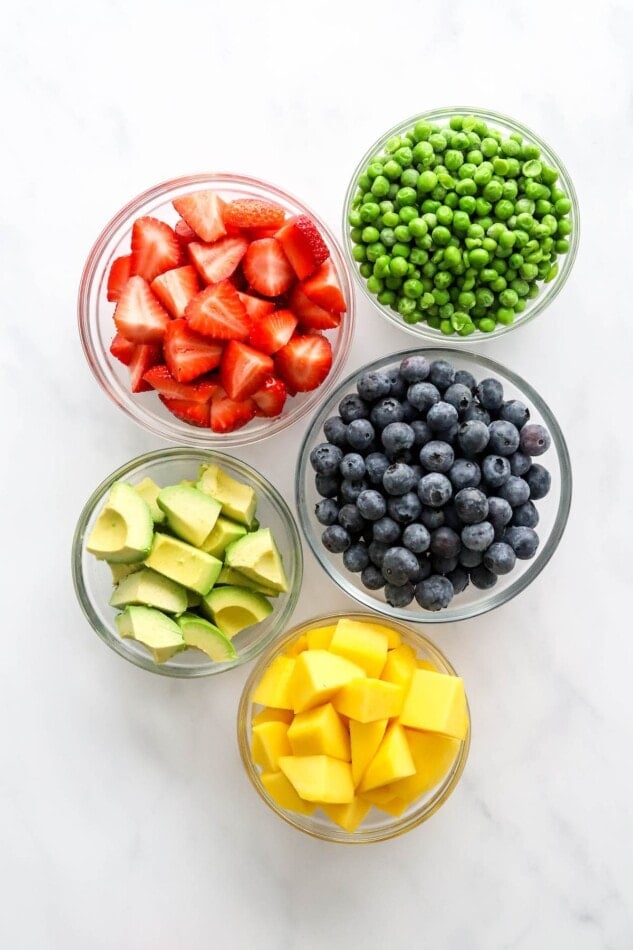 Five glass bowls of various sizes containing: peas, strawberries, blueberries, avocado and mango.