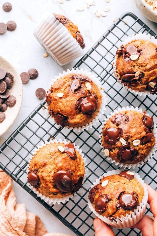 Oatmeal banana muffins on a wire cooling rack. A hand is removing a muffin in the bottom right corner.