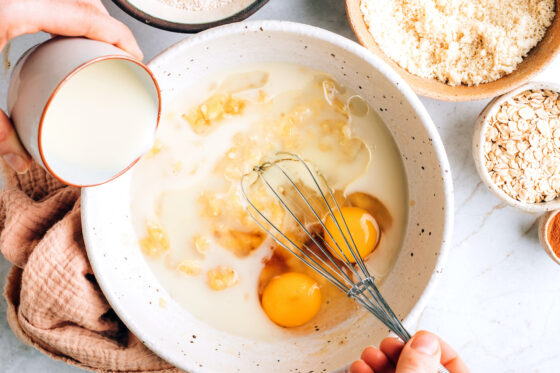 A large mixing bowl with mashed bananas, maple syrup, eggs, vanilla extract, coconut oil and almond milk.