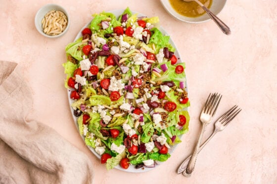 A serving platter with chopped romaine, quinoa, red onion, tomatoes, feta cheese, olives and sliced almonds.