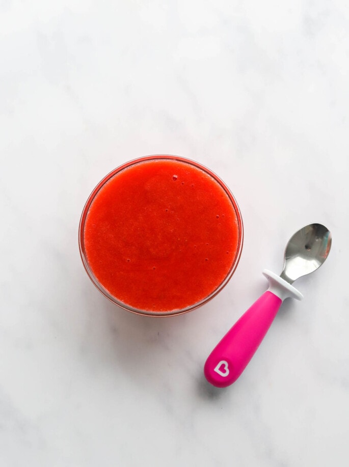 A glass bowl of fresh strawberry puree. A small pink spoon rests next to the bowl.