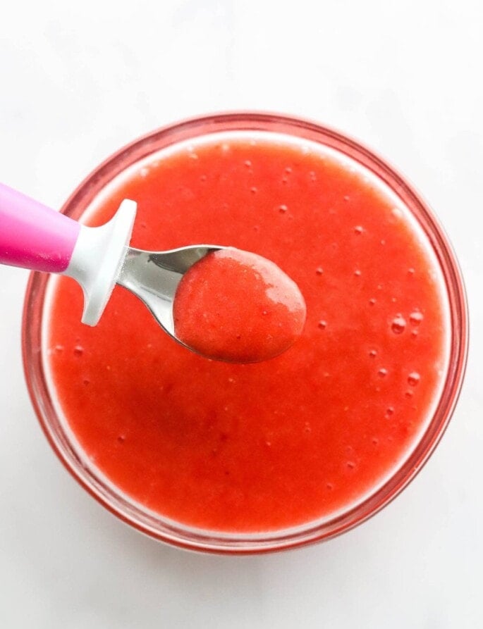 Fresh strawberry puree in a glass bowl. A small spoon is lifting a spoonful out of the bowl.