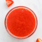 A small glass bowl containing fresh strawberry puree. Strawberry slices are scattered around the bowl.