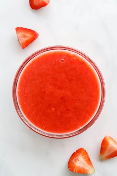 A small glass bowl containing fresh strawberry puree. Strawberry slices are scattered around the bowl.