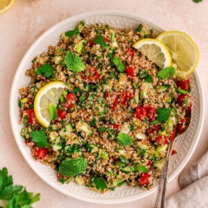 A serving platter with tabbouleh salad topped with fresh mint and lemon slices. A spoon rests on the platter.