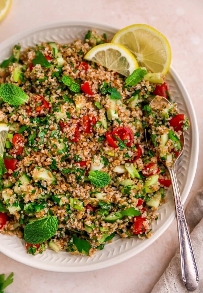 A serving platter with tabbouleh salad topped with fresh mint and lemon slices. A spoon rests on the platter.