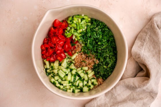 A mixing bowl containing bulgur wheat with parsley, mint, tomatoes, cucumber, green onions, lemon juice, olive oil, sea salt, pepper and allspice added.