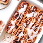 An overhead photo of vegan cinnamon rolls in a baking dish. A roll has been removed from a corner.