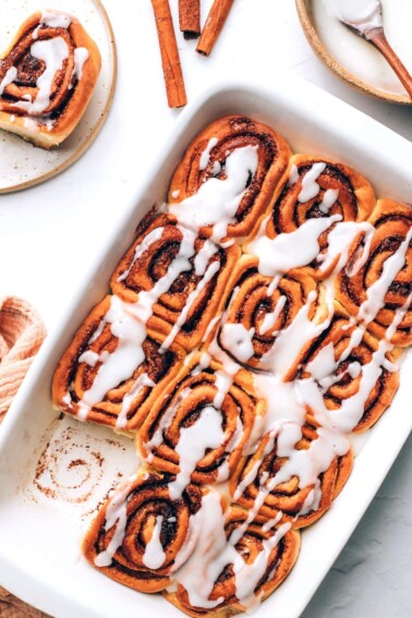 An overhead photo of vegan cinnamon rolls in a baking dish. A roll has been removed from a corner.