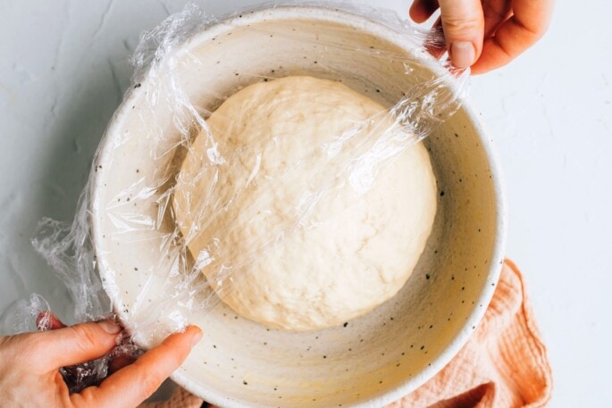 A ball of dough in a bowl being covered with plastic wrap.