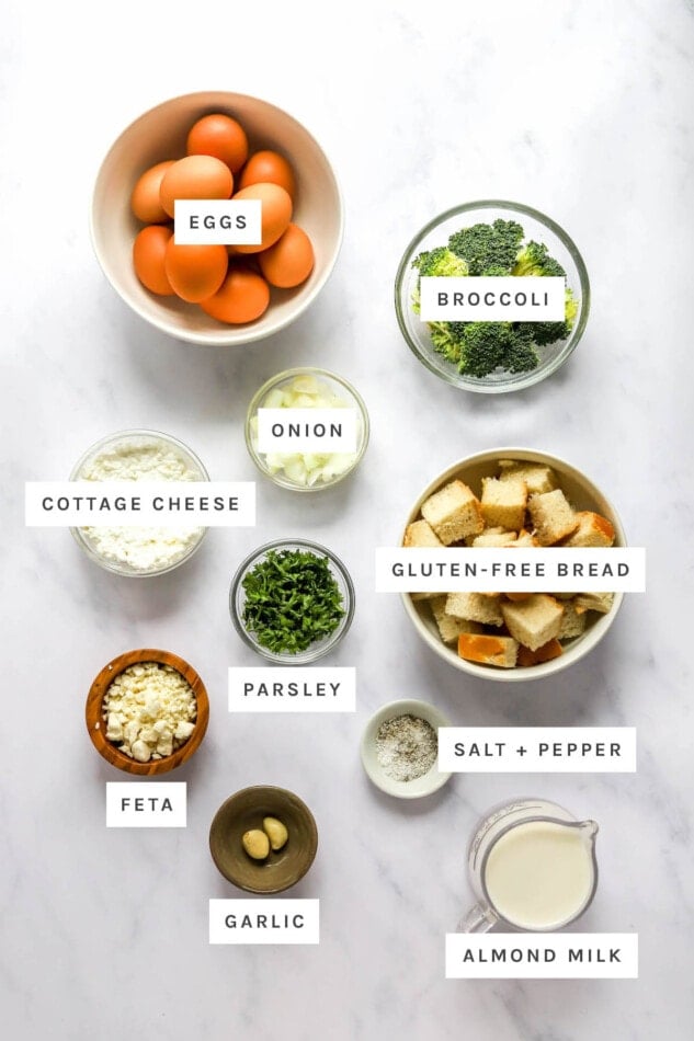 Ingredients measured out to make a healthy breakfast casserole: eggs, broccoli, onion, cottage cheese, gluten-free bread, parsley, feta, garlic, almond milk, salt and pepper.