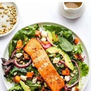 Salmon salad on a plate topped with salmon, red onions, avocado, sweet potatoes, pepitas and feta.