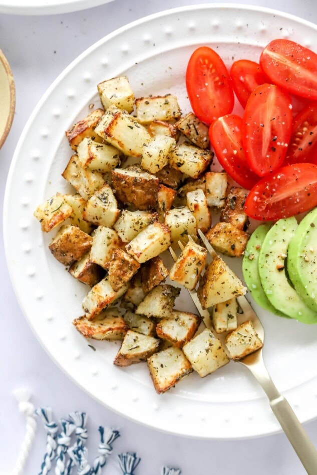 An overhead view of a plate containing air fryer breakfast potatoes alongside tomato and avocado slices. A fork rests in the potatoes.