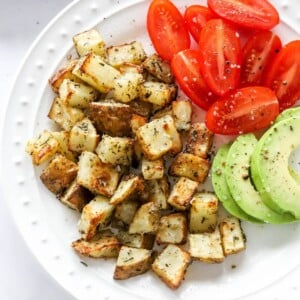 An overhead view of a plate containing air fryer breakfast potatoes alongside tomato and avocado slices.