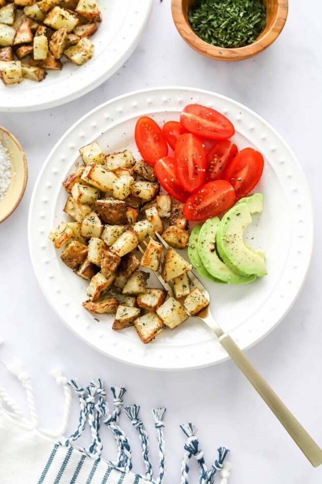 An overhead view of a plate containing air fryer breakfast potatoes alongside tomato and avocado slices. A fork rests in the potatoes.