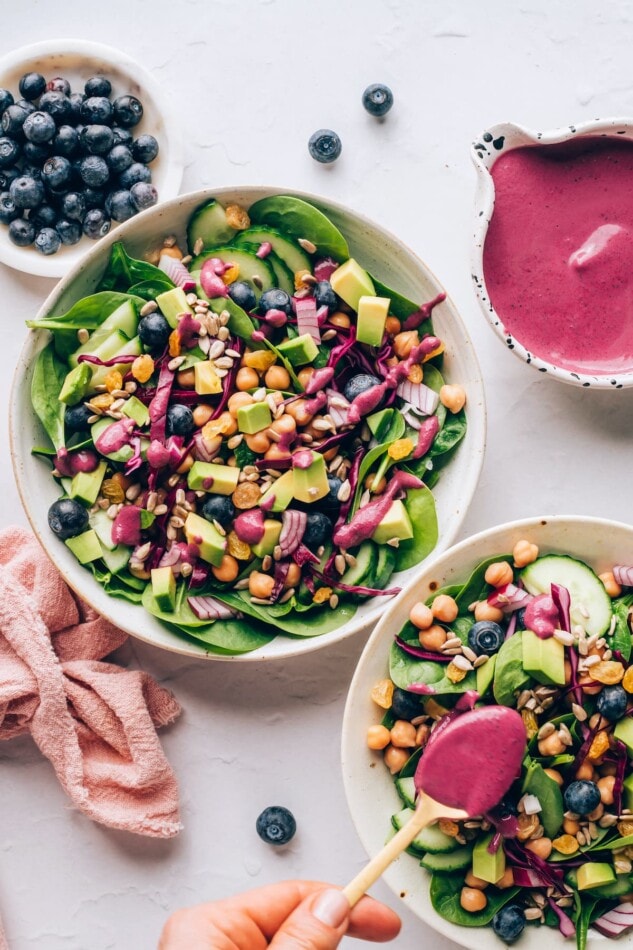 An overhead photo of two bowls of spinach blueberry salad. The salad in the bottom right corner is being drizzled with blueberry tahini dressing.