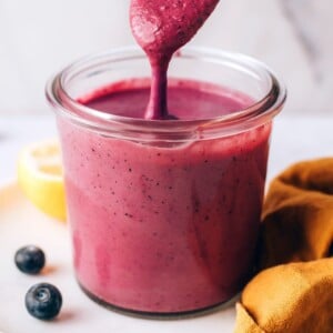 A small jar containing blueberry tahini dressing. A spoon is dripping the dressing back into the jar.