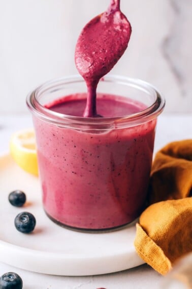 A small jar containing blueberry tahini dressing. A spoon is dripping the dressing back into the jar.