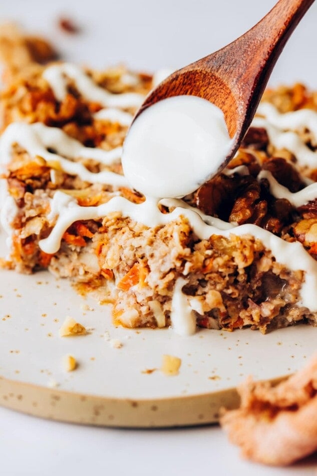 A slice of carrot cake baked oatmeal with a wooden spoon drizzling icing over top.