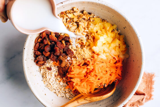 Rolled oats, grated carrots, crushed pineapple, chopped walnuts, raisins, cinnamon, vanilla, flaxseed, maple syrup and a pinch of salt in a mixing bowl. Almond milk is being poured into the bowl.