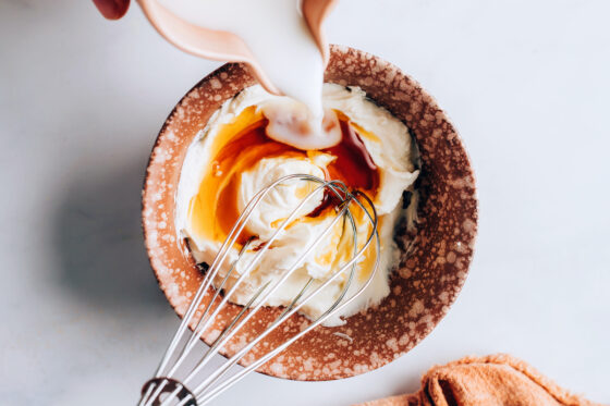 Whipped cream cheese, maple syrup and vanilla in a small bowl. Almond milk is being poured into the bowl.