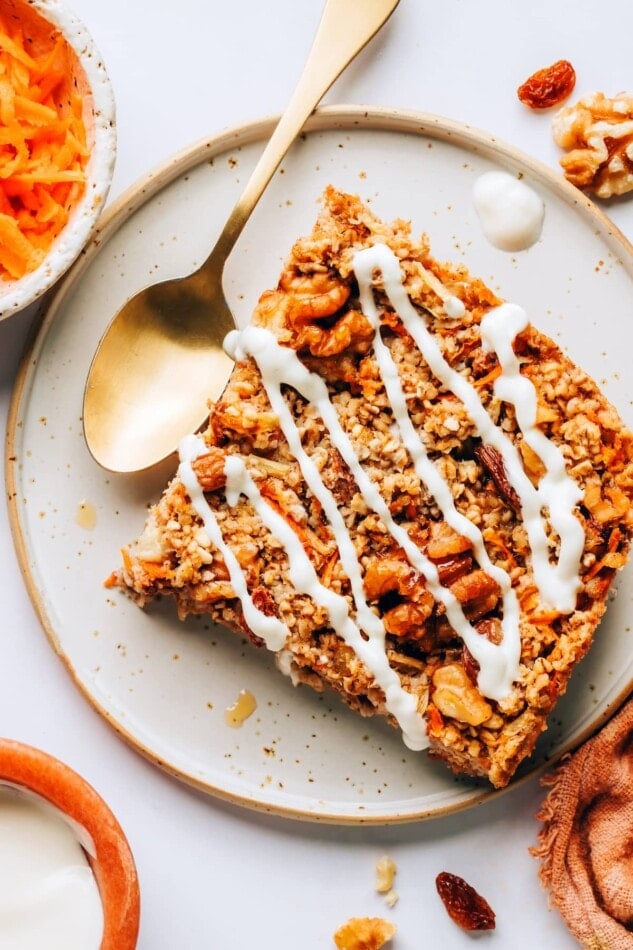 A slice of carrot cake baked oatmeal on a plate. The slice has been drizzled with a maple cream cheese glaze.