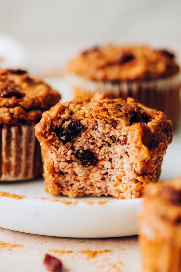 A carrot raisin muffin on a plate. A bite has been taken from the muffin.