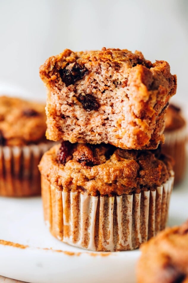 Two muffins stacked on top of each other. The bottom muffin is wrapped in a paper liner, the top muffin has the liner removed with a bite missing.