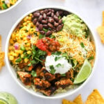 A chicken burrito bowl with corn salad, black beans, guacamole, shredded cheese, lime slices, greek yogurt and salsa served over cilantro lime rice.