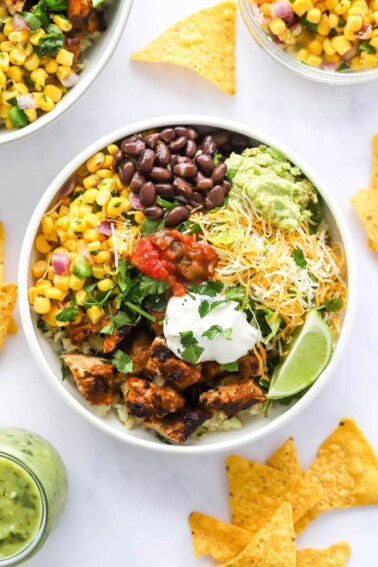 A chicken burrito bowl with corn salad, black beans, guacamole, shredded cheese, lime slices, greek yogurt and salsa served over cilantro lime rice.