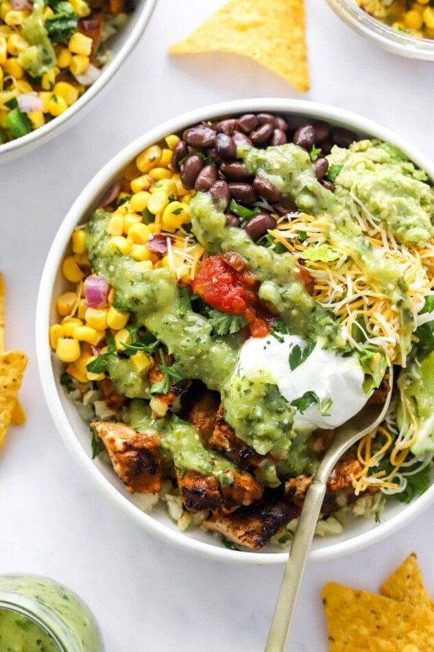 A chicken burrito bowl with corn salad, black beans, guacamole, shredded cheese, lime slices, greek yogurt and salsa served over cilantro lime rice. It has been drizzled with a tomatillo avocado dressing.