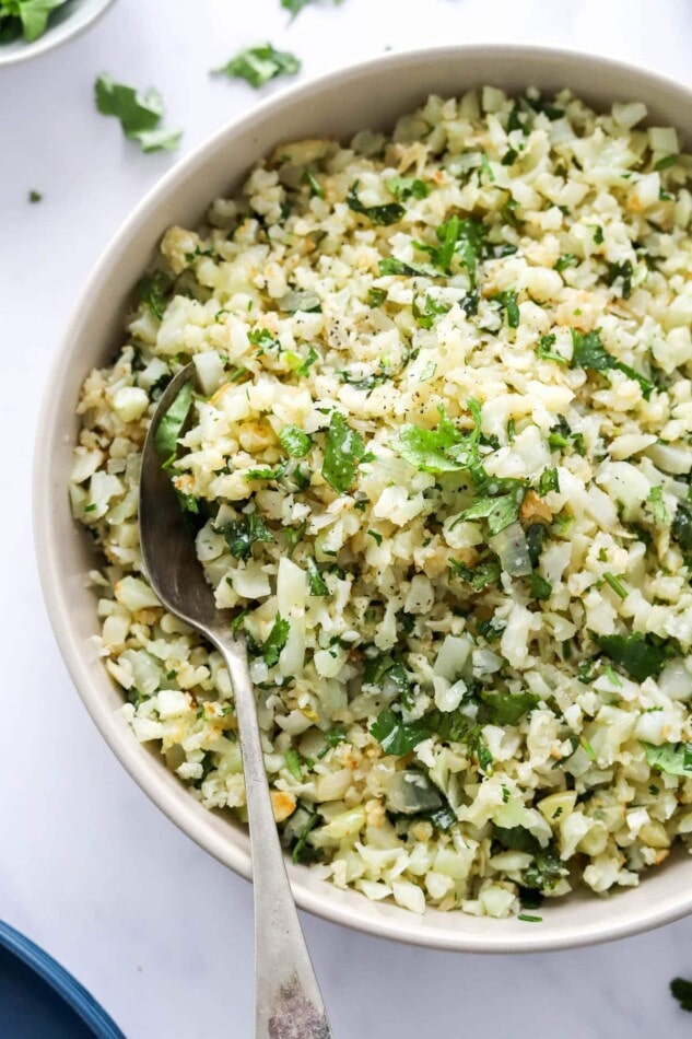 A closeup of a serving bowl containing cilantro lime cauliflower rice. A spoon rests in the bowl.