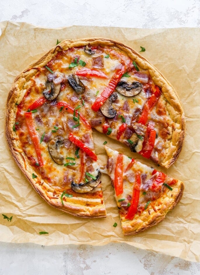 An overhead view of cloud bread pizza on a sheet of brown parchment paper. A slice has been cut and slightly removed from the pizza.