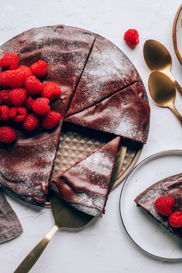 An overhead view of a flourless chocolate cake topped with raspberries and powdered sugar. A slice of cake is resting on a serving utensil.