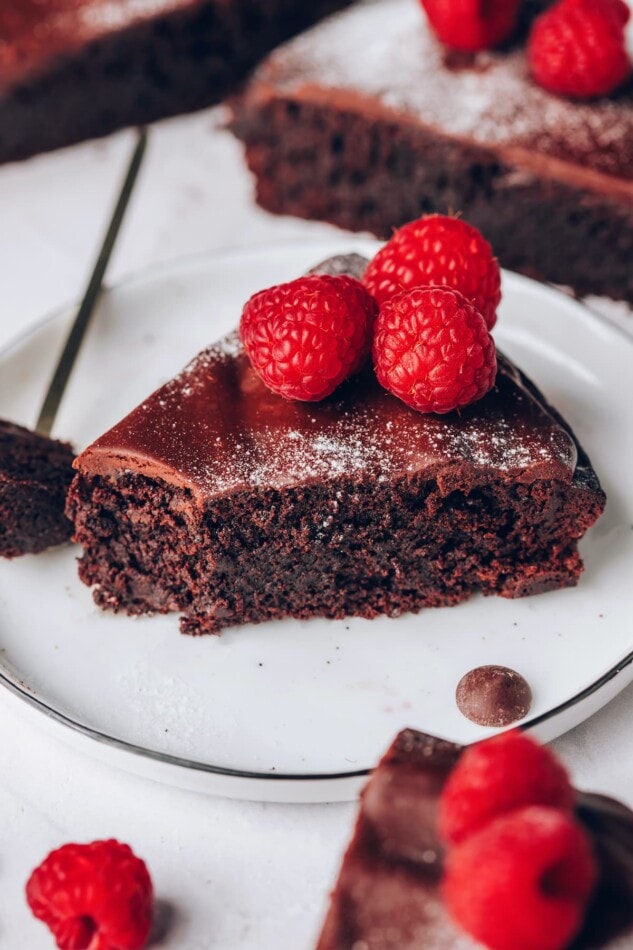 A slice of flourless chocolate cake, sprinkled with powdered sugar and topped with raspberries on a plate. A bite has been taken out of the slice.