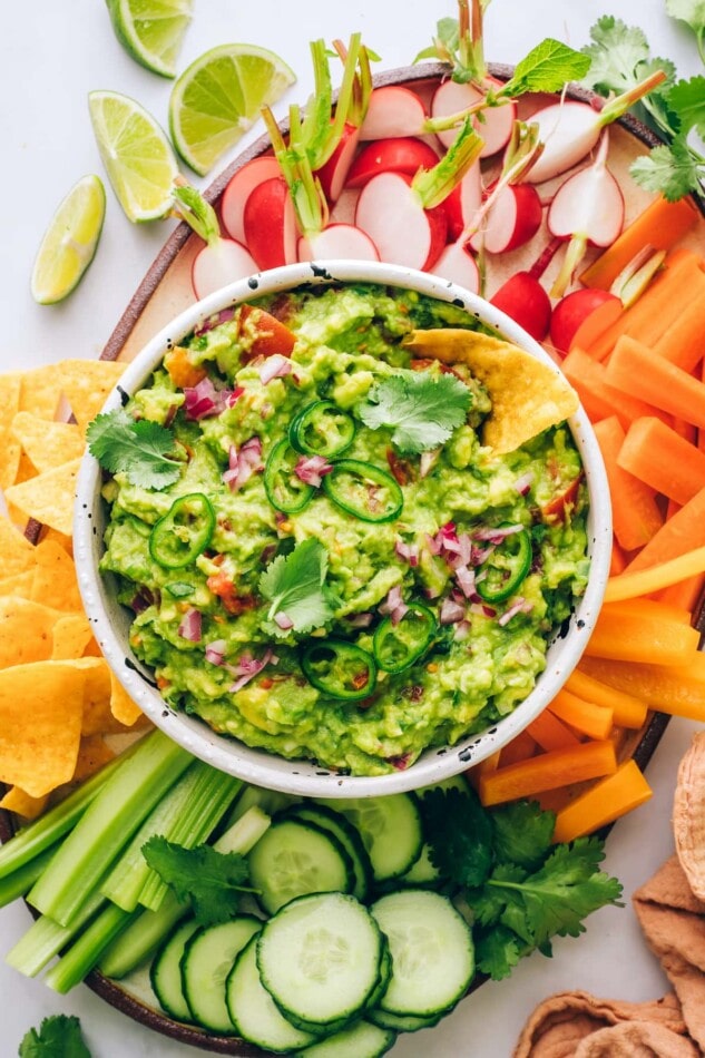 A serving bowl of guacamole surrounded by carrots, radishes, chips, celery, cucumber slices, orange pepper and carrots for dipping.