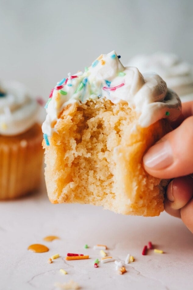 A healthy vanilla cupcake with the liner removed held up by a hand, with a bite taken out of it.